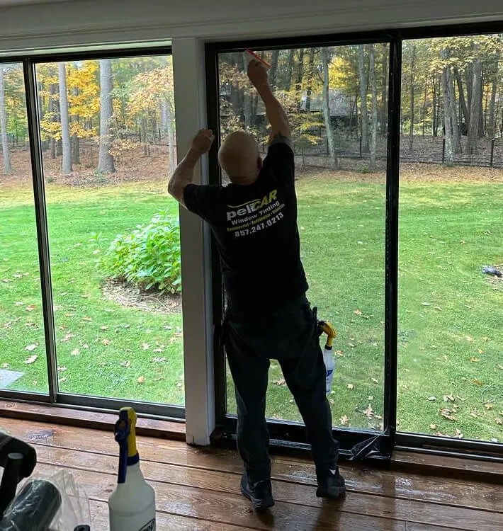 A professional window tinter from peliCAR Window Tinting installing a tint film on a large sliding glass door. The installer, wearing a branded shirt, is carefully applying the film while a spray bottle and tools are nearby. The background shows a lush green yard with trees in autumn colors.