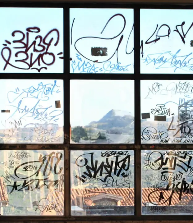A large window covered in various graffiti tags in different colors and styles. The glass panes are filled with overlapping words, symbols, and scribbles, partially obscuring a scenic view of mountains and rooftops in the background.