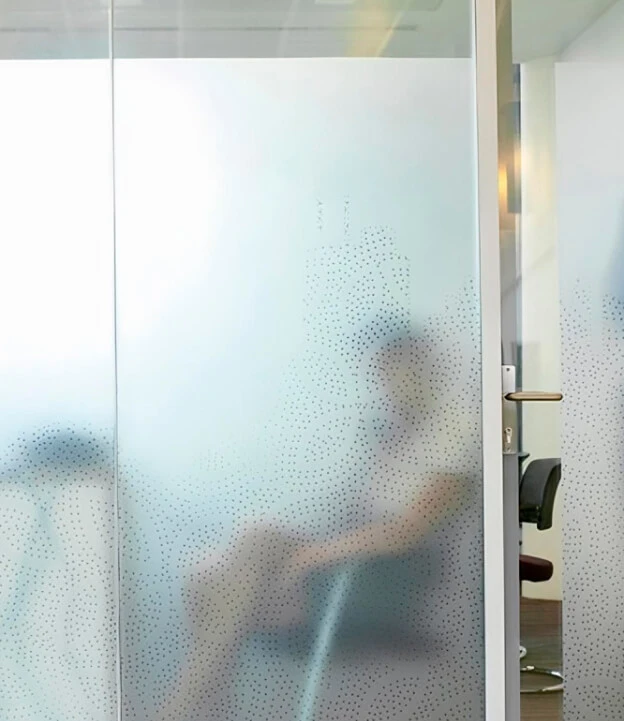A frosted glass office partition with a decorative dotted pattern providing privacy. A blurred silhouette of a seated person is visible behind the glass, creating a modern and professional ambiance.