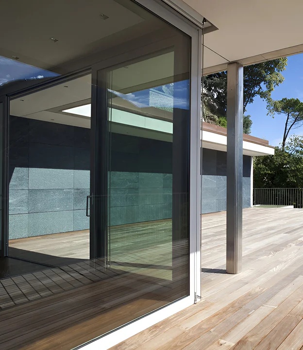 A modern sliding glass door with clear glass, reflecting the outdoor surroundings and wooden floor, leading to a patio.