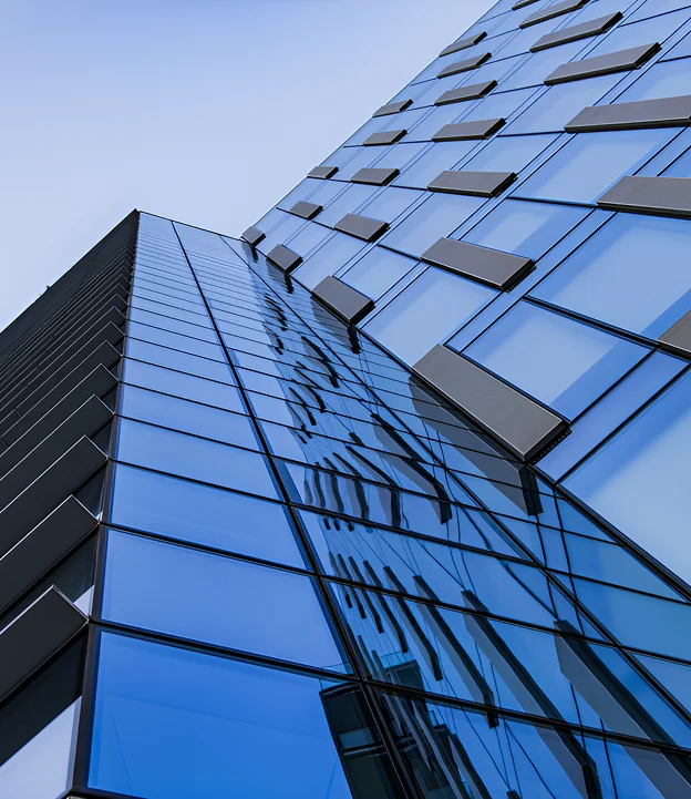 A modern glass building with reflective blue-tinted windows captured from a low angle. The facade features geometric protrusions that add depth and contrast against the clear sky.