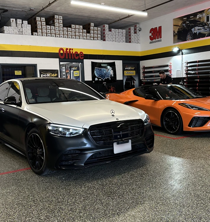 "A sleek two-tone Mercedes-Benz with a black and white wrap sits next to an orange Chevrolet Corvette inside a professional tinting and wrapping shop. The workshop has a '3M' sign, an 'Office' sign, and neatly stacked tint film rolls on the walls. A technician is seen working in the background, indicating an active workspace.