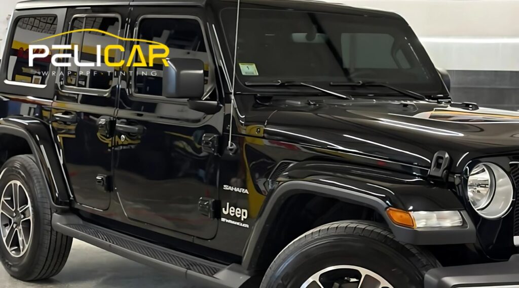 A shiny black Jeep Wrangler Sahara with tinted windows parked inside an auto detailing shop. The shop's logo, "PELICAR Wrap · PPF · Tinting," is reflected on the vehicle's side, showcasing the high gloss of the tint and wrap finish.