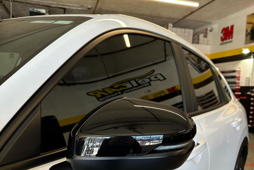 Close-up of a white car with freshly tinted windows and a glossy black side mirror, parked inside a tinting studio. The window tint appears dark and sleek, reflecting the Pelicar branding and shop interior in the glass.