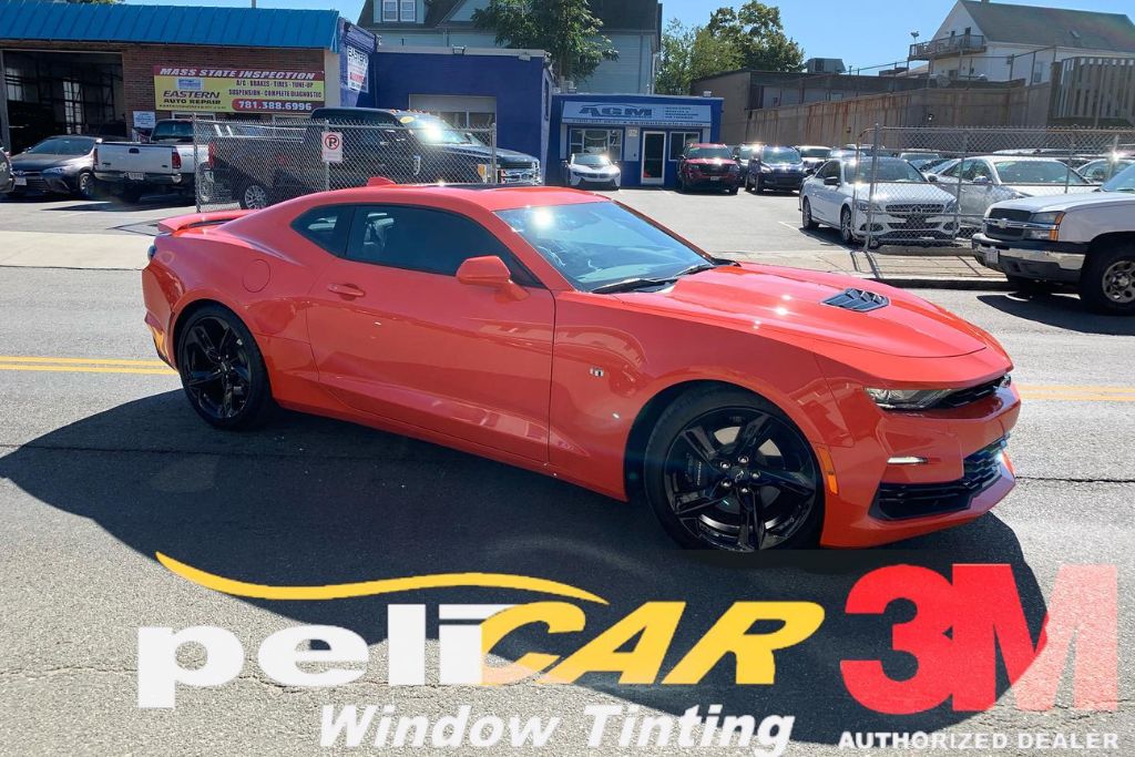 A bright orange sports car with tinted windows is parked on a street outside an auto shop, featuring large logos for Pelicar Window Tinting and 3M Authorized Dealer overlaid at the bottom.