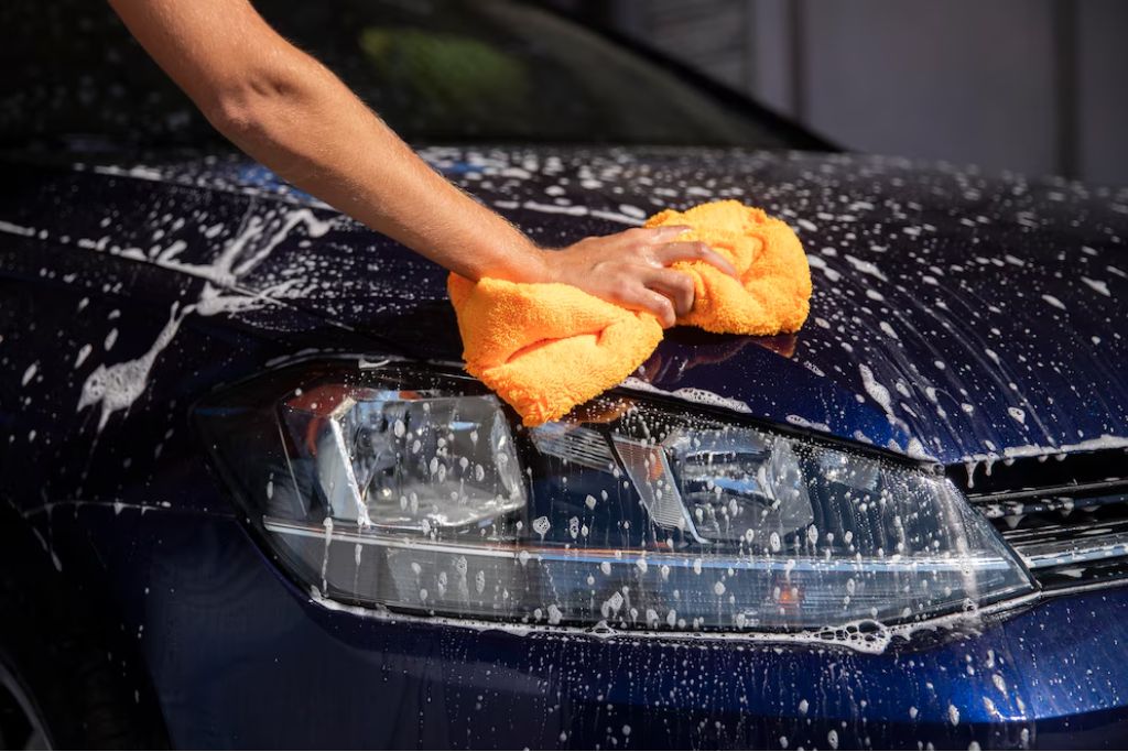 A close-up of a person washing a dark blue car by hand with a bright orange microfiber cloth and foamy soap, emphasizing careful maintenance for a vinyl wrap.
