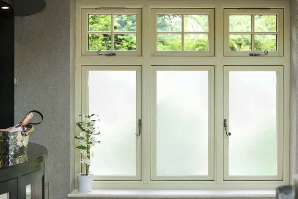 A set of classic-style windows with the lower panes covered in frosted privacy film, allowing natural light to filter through while obscuring the view from both sides. The upper panes remain clear, offering an open view of the greenery outside.