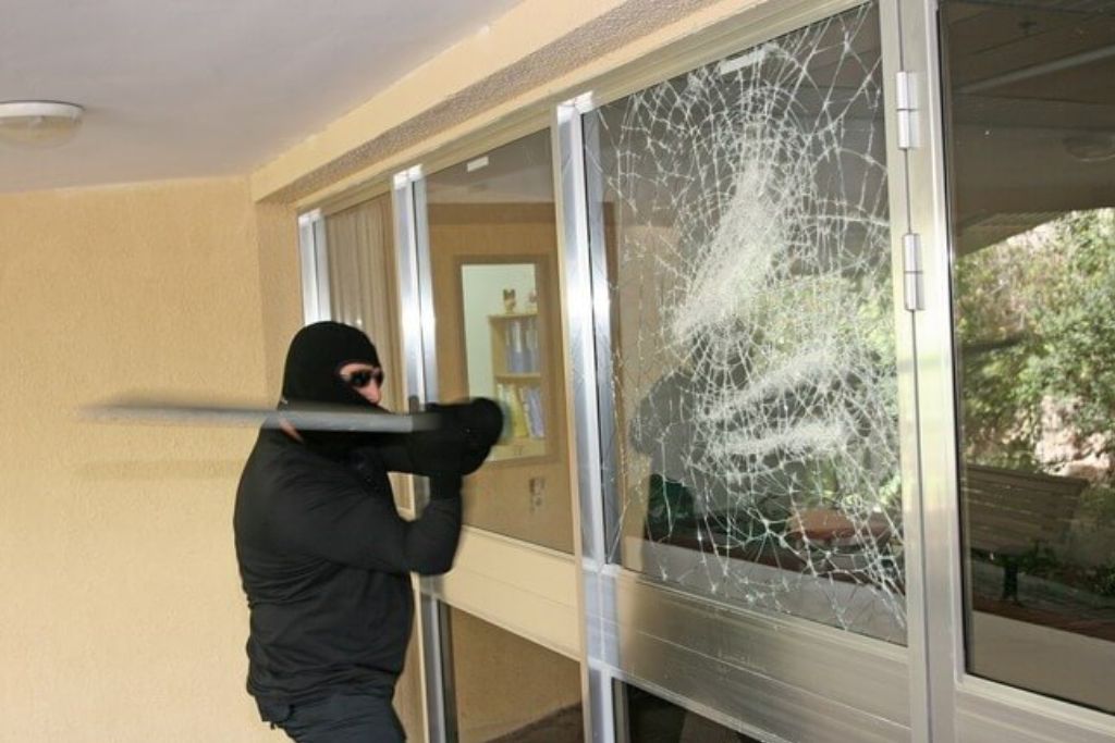 A person in dark clothing and a ski mask strikes a large glass door with a metal object. The glass is heavily cracked in a spiderweb pattern but remains intact, showing the protective effect of a security window film.