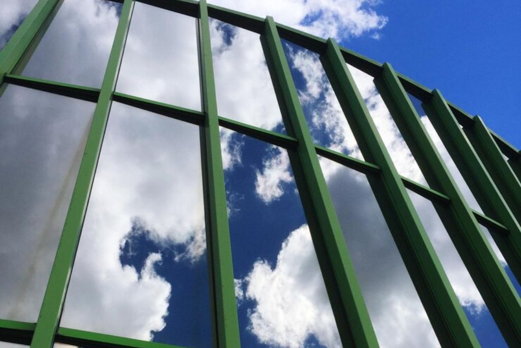 A close-up view of a modern glass building with green framing, reflecting bright blue sky and white clouds. The windows have a mirrored tint, suggesting the use of solar control film to reduce glare and heat.