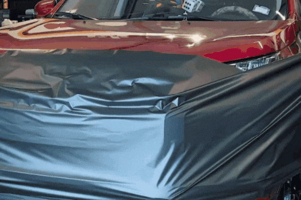 A time-lapse GIF showing a vinyl wrap being applied to the hood of a red car, with the black film smoothly laid over the surface by hand.