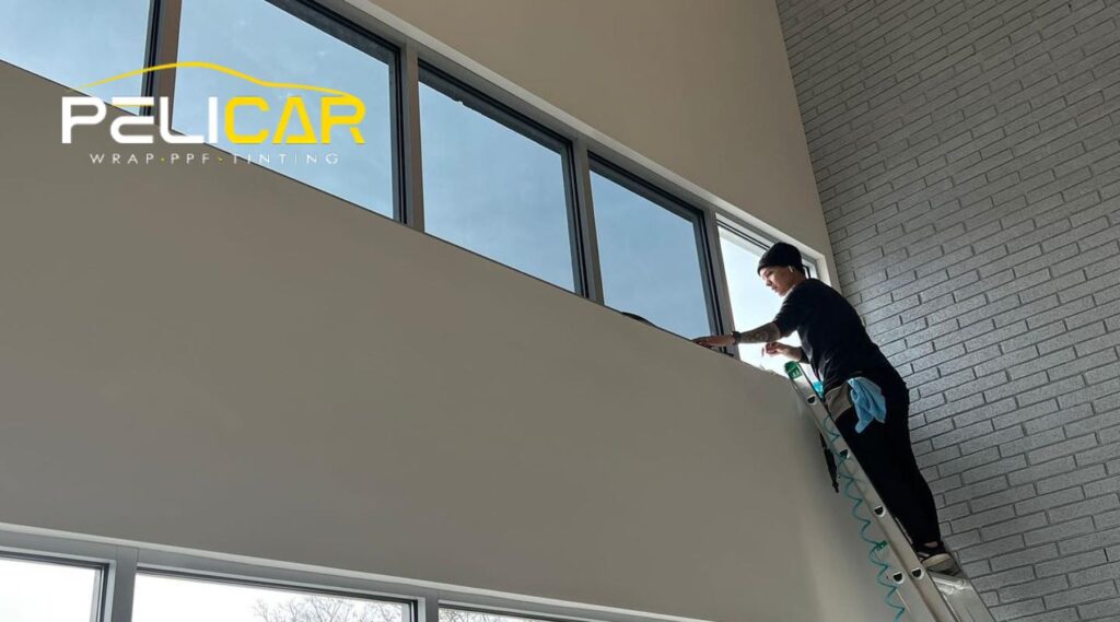 A technician from Pelicar Window Tinting stands on a ladder inside a modern home, applying window film to large, upper-level windows. The person is focused on the work, using tools and wearing casual black clothing with a cleaning cloth tucked into their waistband.