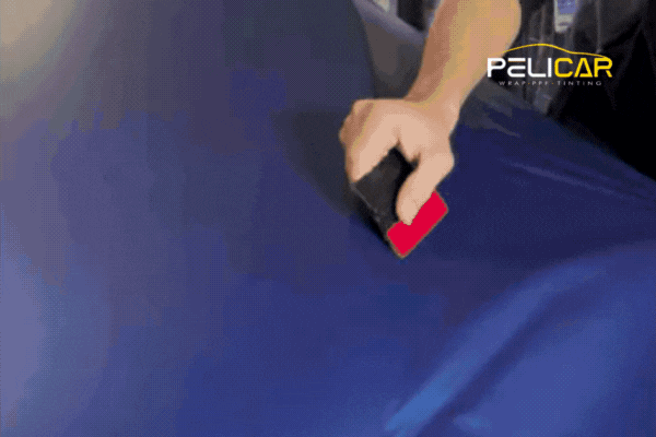A technician smooths out a deep blue vinyl wrap across a car’s hood using a squeegee tool, with the Pelicar logo visible in the corner of the screen.