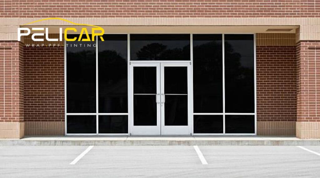 A commercial storefront with large, dark-tinted glass windows and double glass doors, framed by a red brick building. The Pelicar logo is overlaid in the top-left corner, representing their expertise in window tinting solutions.