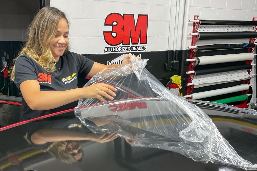 A smiling technician wearing a 3M Authorized Dealer shirt applies a clear paint protection film to the hood of a black car in a workshop, with a rack of vinyl rolls and a prominent 3M logo on the wall behind her.