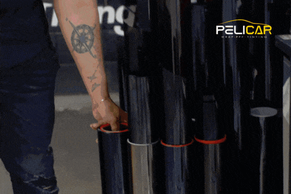 A tattooed arm selects a roll of window tint film from a vertical display of glossy black film rolls inside a professional tinting shop. The Pelicar Window Tinting logo appears in the top-right corner.