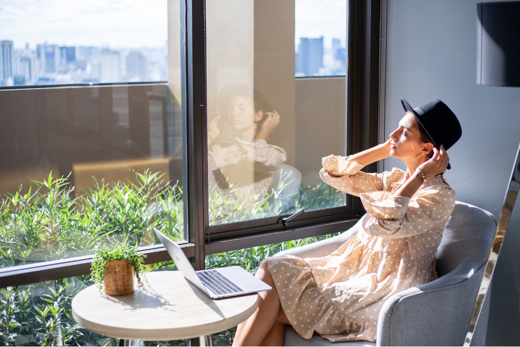 A woman sits comfortably in a sunlit room near tinted windows, with a laptop on the table and greenery outside. The window tint reduces glare and harmful UV exposure, demonstrating the benefits of UV protection for interiors and employees.