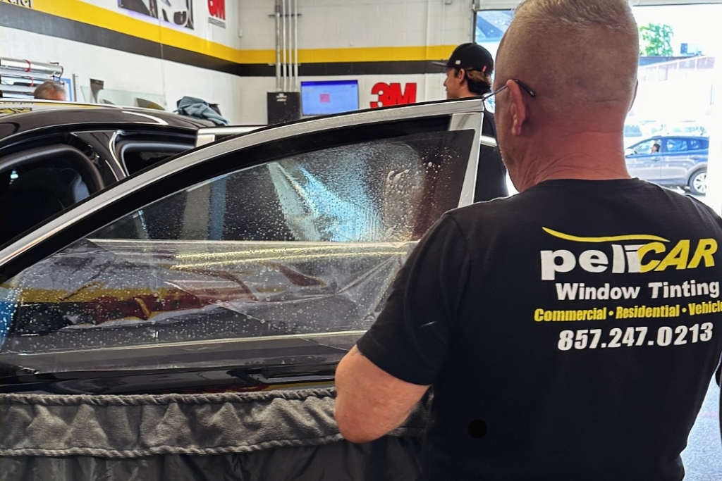 A technician at Pelicar Window Tinting in Malden applies tint film to a car's side window using a squeegee in a clean, well-lit garage. The worker wears a black branded t-shirt and works with precision, highlighting professional service during the fall season.