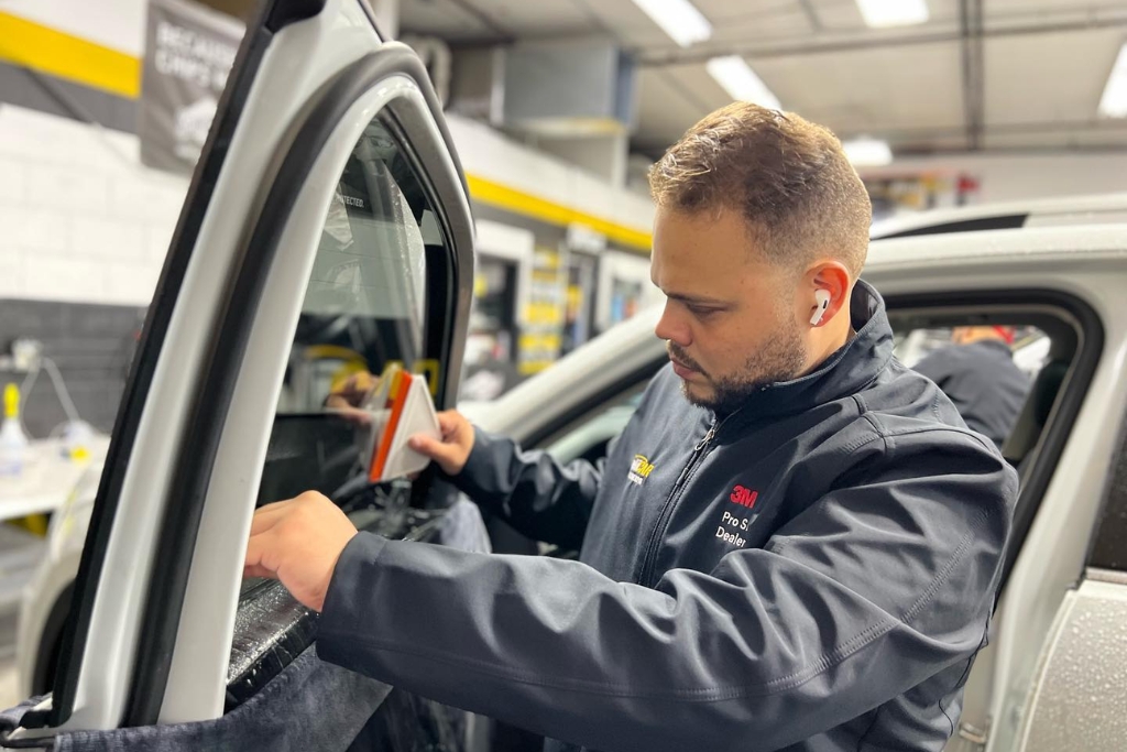 A technician at Pelicar in Malden carefully applies window tint film to a car’s side window using a squeegee tool, wearing a branded 3M jacket and working in a clean, well-lit shop. The image reinforces that fall is a smart time to tint your car windows thanks to increased appointment availability and seasonal deals.