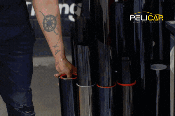 A tattooed technician at Pelicar in Malden, MA selects a roll of Paint Protection Film from a neatly organized display of vinyl and PPF materials. This image supports the topic Choosing the Right Installer in Malden, MA by showcasing professional-grade tools and a detail-focused shop environment.