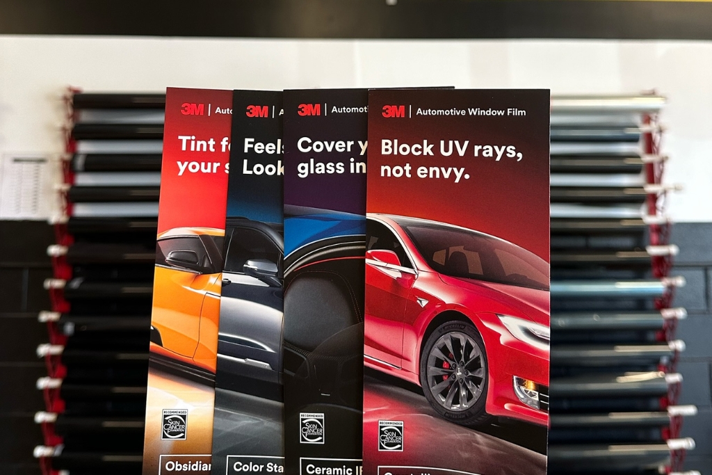 Four 3M automotive window film brochures are fanned out in front of a wall of tint rolls at Pelicar in Malden, each promoting a different tint series—Obsidian, Color Stable, Ceramic IR, and Crystalline—with bold colors and phrases like "Block UV rays, not envy." The image illustrates the variety of options available during a smart time to tint your car windows in fall.