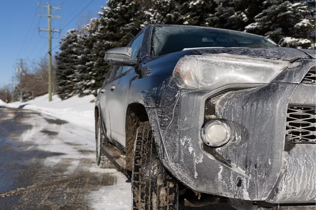 A close-up of a dirty SUV in Malden, Massachusetts, coated with layers of salt, slush, and grime on a snowy roadside, showing real-world effects of harsh winter conditions. The image illustrates How Winter Damages Paint through chemical buildup, road debris, and moisture exposure.
