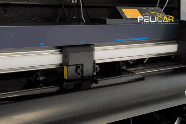 Close-up of a precision cutting machine at Pelicar in Malden slicing black window tint film, with the Pelicar logo visible in the corner. This process highlights expert craftsmanship and why fall is a smart time to tint your car windows with professional-grade materials and equipment.