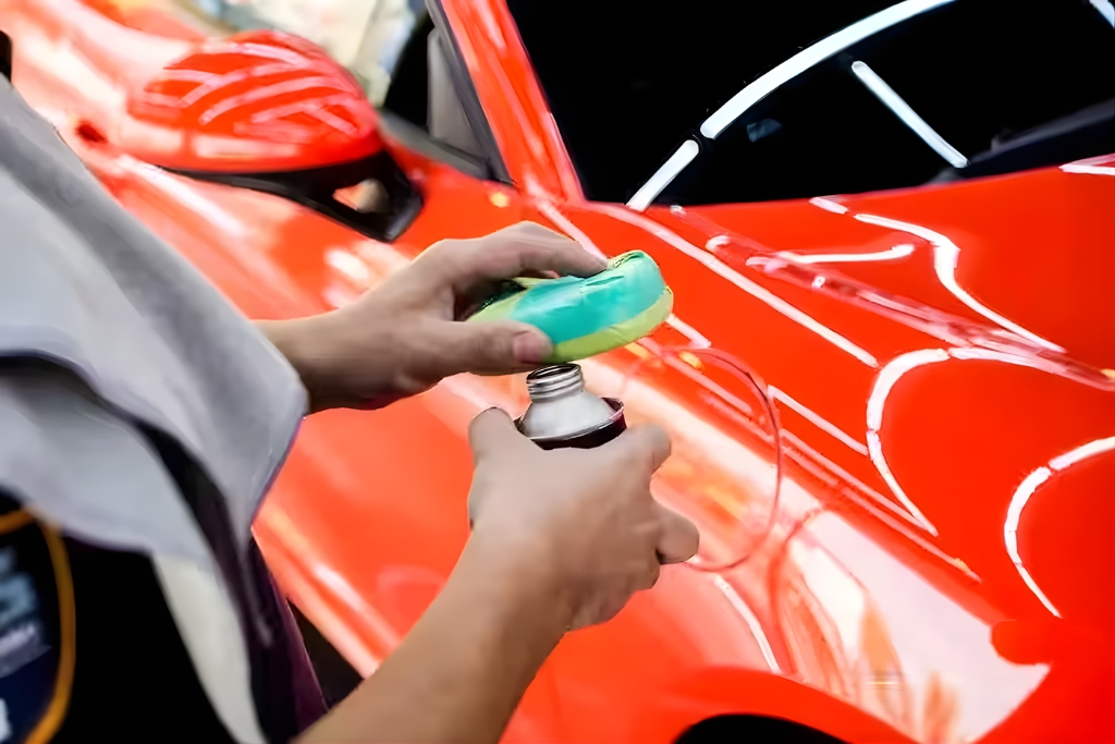 A detailer applies wax to a polishing pad beside a glossy red sports car, highlighting a common paint protection method. The image contrasts traditional waxing with more durable options like Paint Protection Film in the context of PPF vs. Other Winter Paint Protection Options.