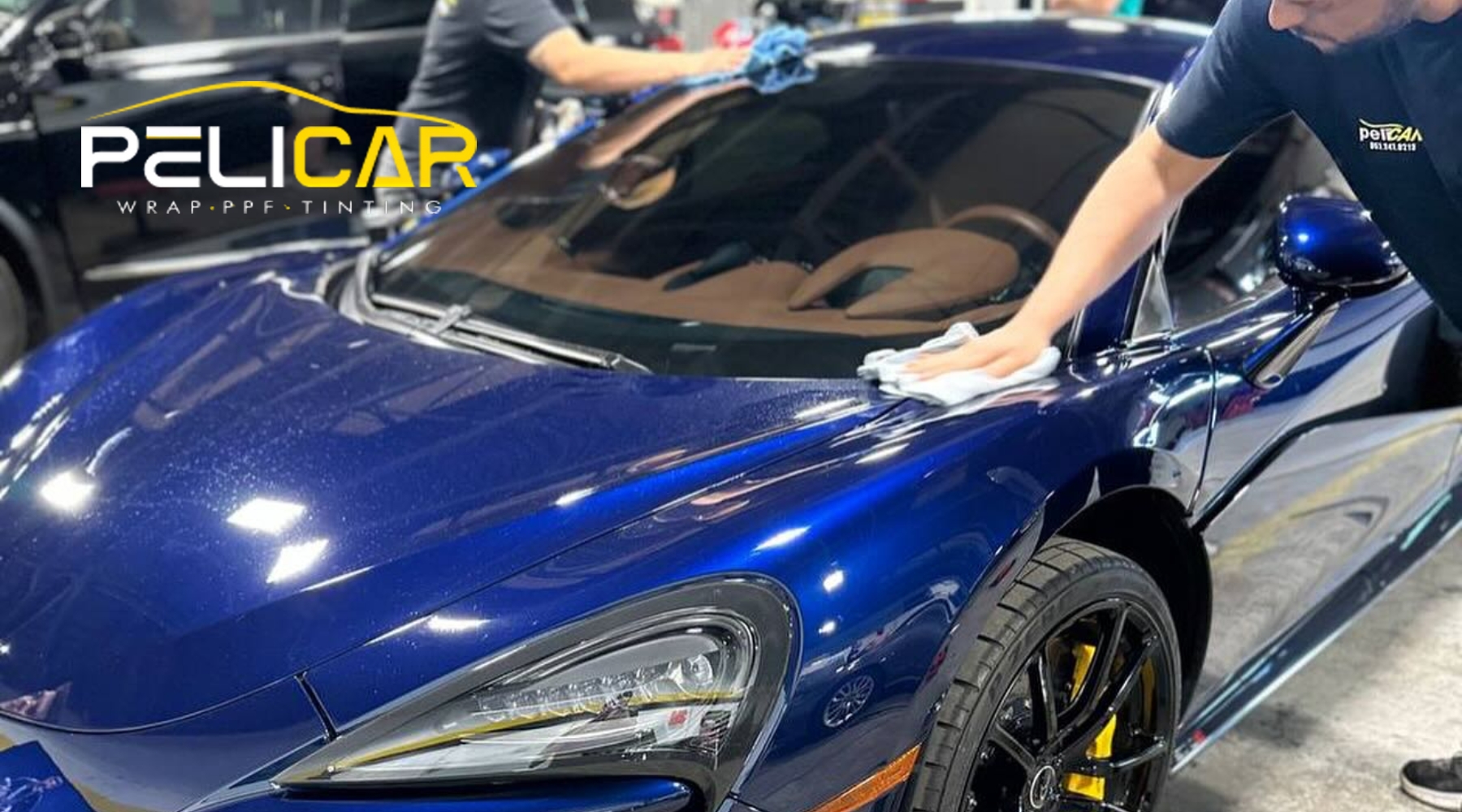 A technician in Malden, Massachusetts carefully hand-polishing a deep blue sports car inside a detailing shop, prepping the surface before applying Paint Protection Film. The image supports the topic "Does PPF Help" by showing the paintwork being cleaned before protective treatment for winter.