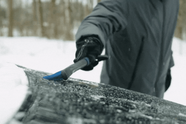 A person in winter gear scrapes ice off a snow-covered car windshield, illustrating the wear and tear vehicles endure in cold climates. This scene supports the topic Benefits of PPF in Winter by highlighting how Paint Protection Film reduces the need for aggressive scraping and shields the paint underneath.