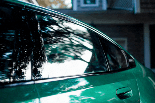 Close-up of a green car's tinted rear side window reflecting surrounding trees and a house, showcasing a smooth, bubble-free finish achieved during fall in Malden. The image supports why fall is a smart time to tint your car windows due to ideal curing conditions.