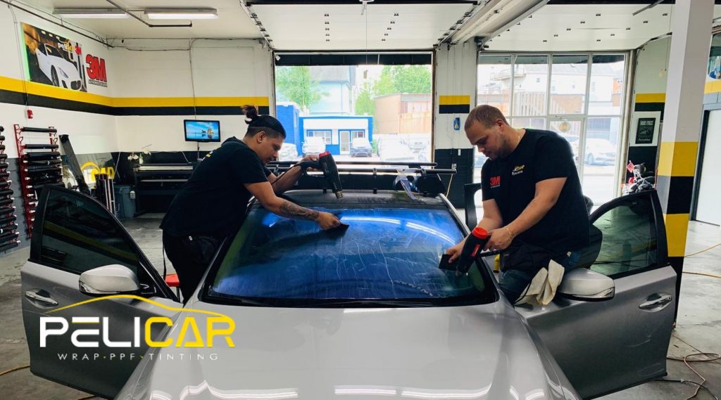 Two technicians at Pelicar in Malden apply window tint to a silver car’s front windshield using heat guns and squeegees inside a well-lit shop. The image highlights why fall is a smart time to tint your car windows, showing optimal working conditions and professional application.