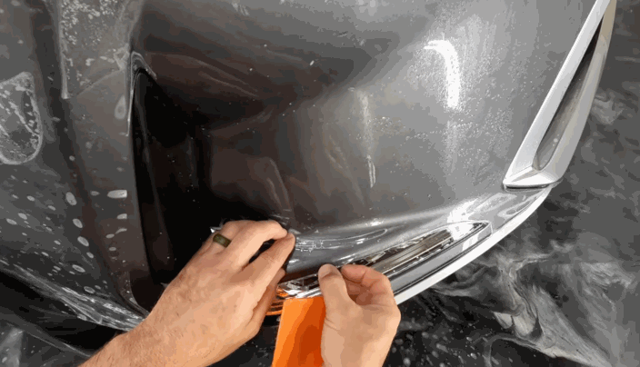 Hands using an orange squeegee to precisely apply paint protection film around a curved bumper edge on a gray vehicle.