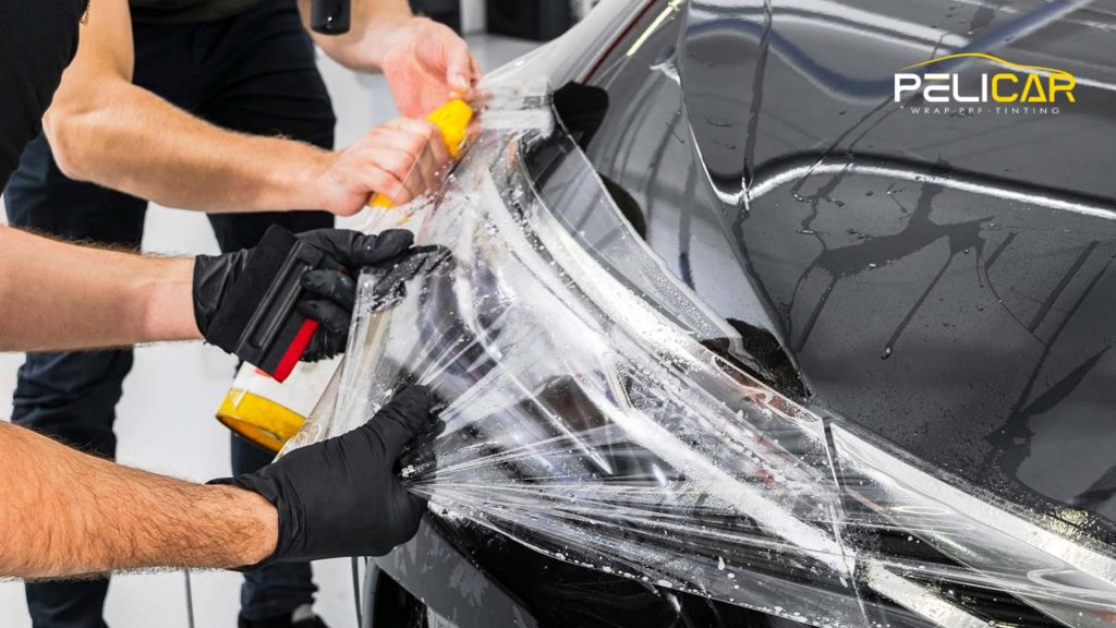 Close-up of two people installing paint protection film on the front headlight and hood of a black car using squeegees and gloves, with the Pelicar logo in the corner.