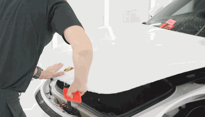A technician smooths out clear paint protection film over the hood of a white Porsche using a red squeegee.