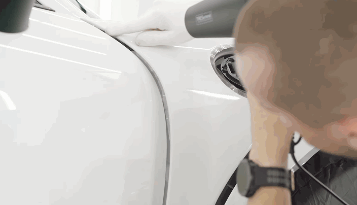 Close-up of a technician using a heat gun to secure paint protection film around the curved edge of a white car's fender.