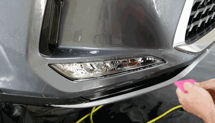 Close-up of a technician applying clear paint protection film to a gray car’s front bumper using a pink squeegee, smoothing out water and air bubbles.