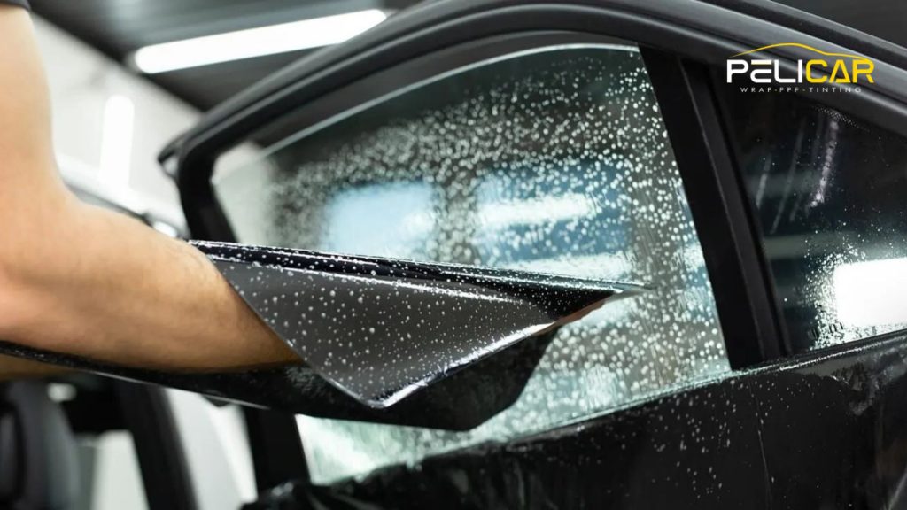 Close-up of a technician applying window tint film to a wet car window, showing water droplets on the glass and a partially smoothed tint sheet.