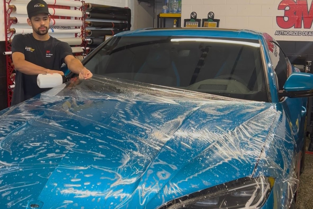A Pelicar technician in Malden, MA smooths Paint Protection Film over a blue car’s hood, with water droplets visible on the surface, showing the detailed process of Prepping Your Car’s Paint Before Winter to prevent seasonal damage.