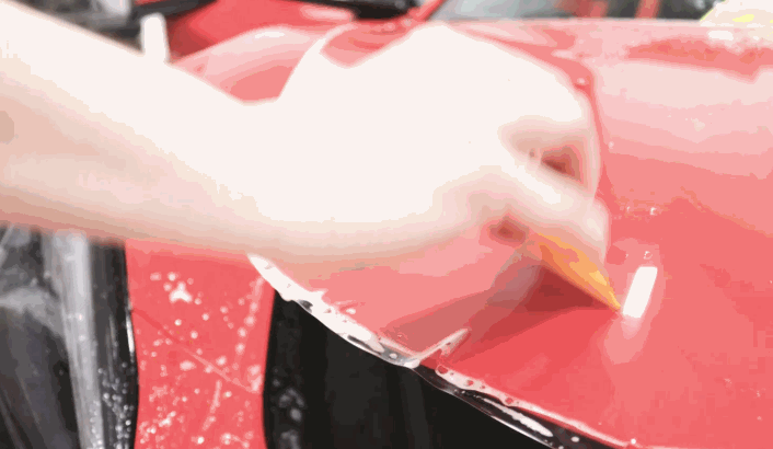 A person uses a squeegee to smooth out a clear film on the hood of a red car during a PPF installation.