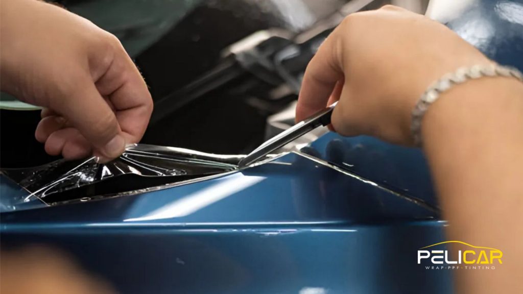 Close-up of hands trimming excess paint protection film from a blue vehicle using a precision blade.