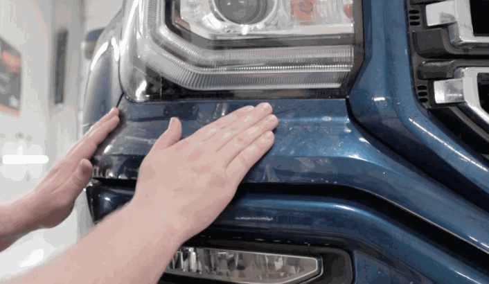 Hands smoothing a paint protection film onto the front bumper of a blue truck near the headlight.