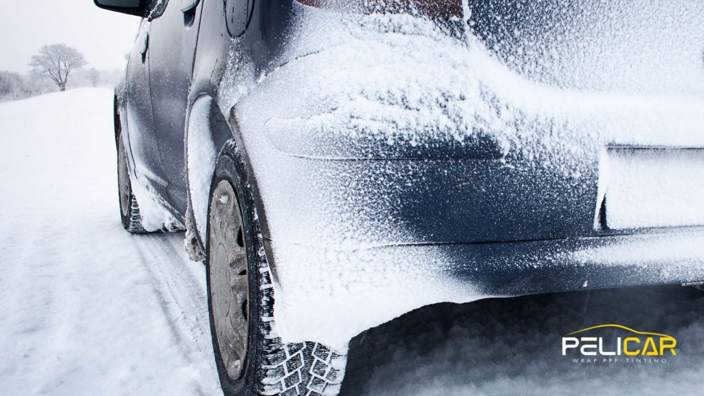 Rear view of a dark vehicle covered in snow and ice on a winter road, highlighting the buildup around the wheel and bumper.