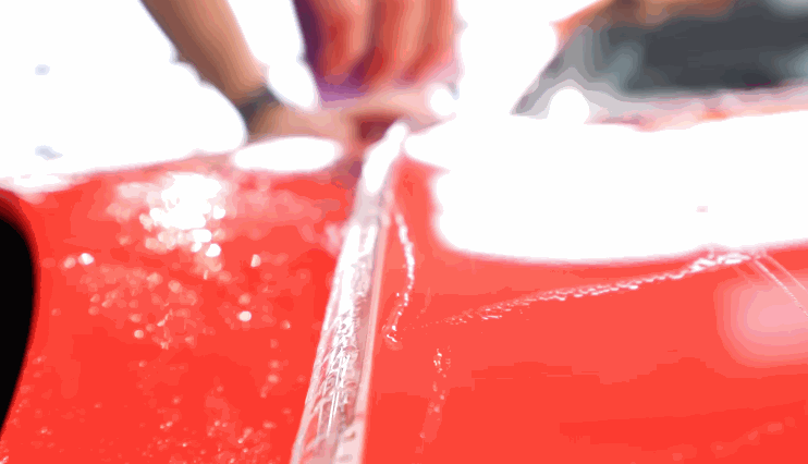 Close-up of hands applying clear paint protection film (PPF) to a glossy red vehicle surface, with water and soap solution visible during installation.