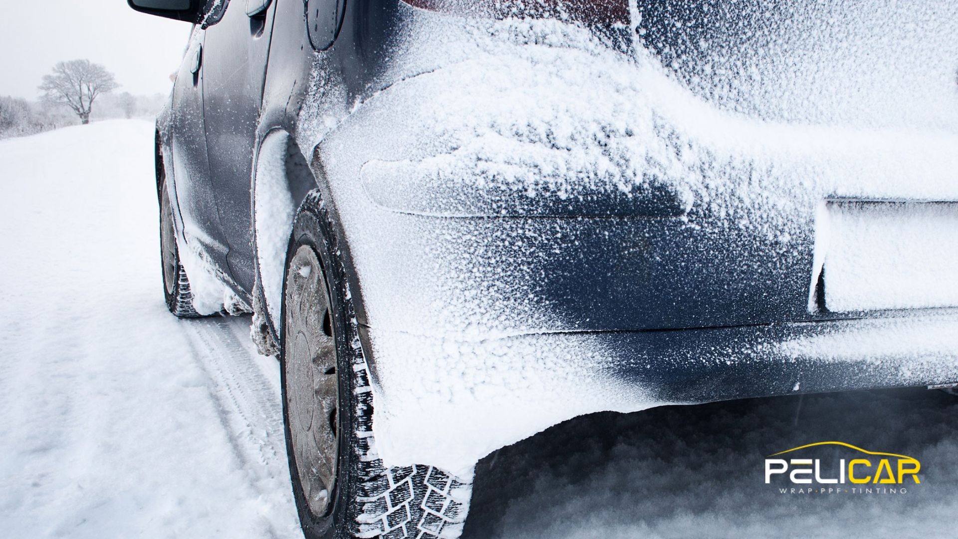 Rear view of a dark vehicle covered in snow and ice on a winter road, highlighting the buildup around the wheel and bumper.