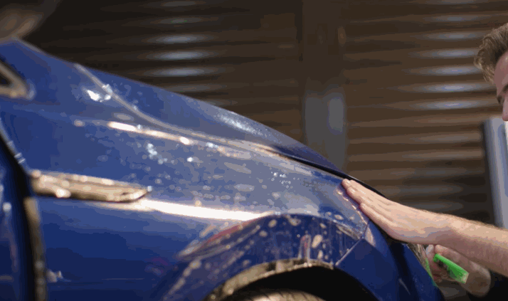 A man applies blue paint protection film to a car fender, using his hand to press it into place. The car is wet, suggesting the use of a slip solution to aid in the installation.