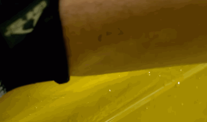 Close-up of a person applying a yellow-tinted paint protection film onto a car’s surface, with a black-gloved hand pressing the film down smoothly.