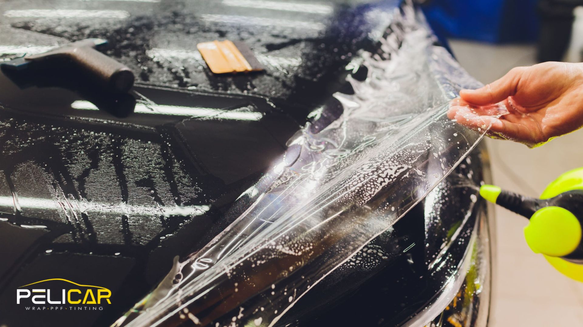 Transparent paint protection film being pulled over the wet surface of a black car hood, with installation tools and water droplets visible. The installer’s hand stretches the film while a spray bottle sits nearby.
