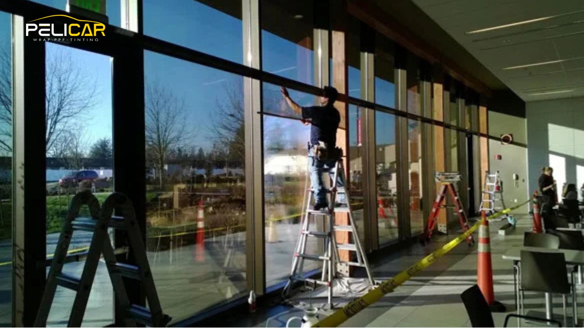 Installer on a ladder applies commercial window tint to tall glass panels inside a modern building, surrounded by caution tape and tools.