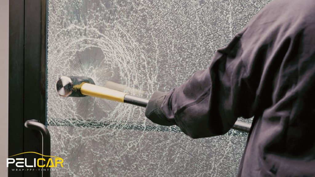 Close-up of a sledgehammer pressed against a cracked glass door that remains held together by security window film, with the Pelicar logo in the corner.