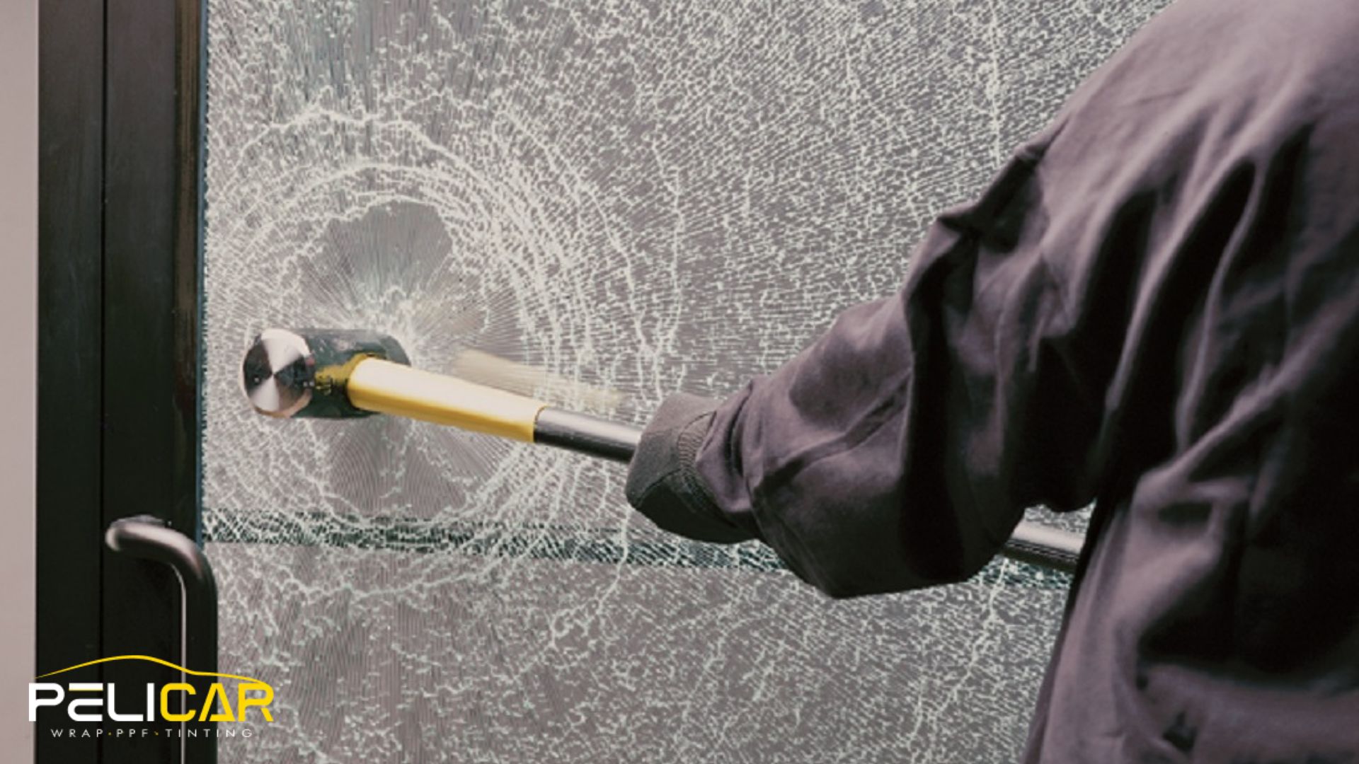 Close-up of a sledgehammer pressed against a cracked glass door that remains held together by security window film, with the Pelicar logo in the corner.